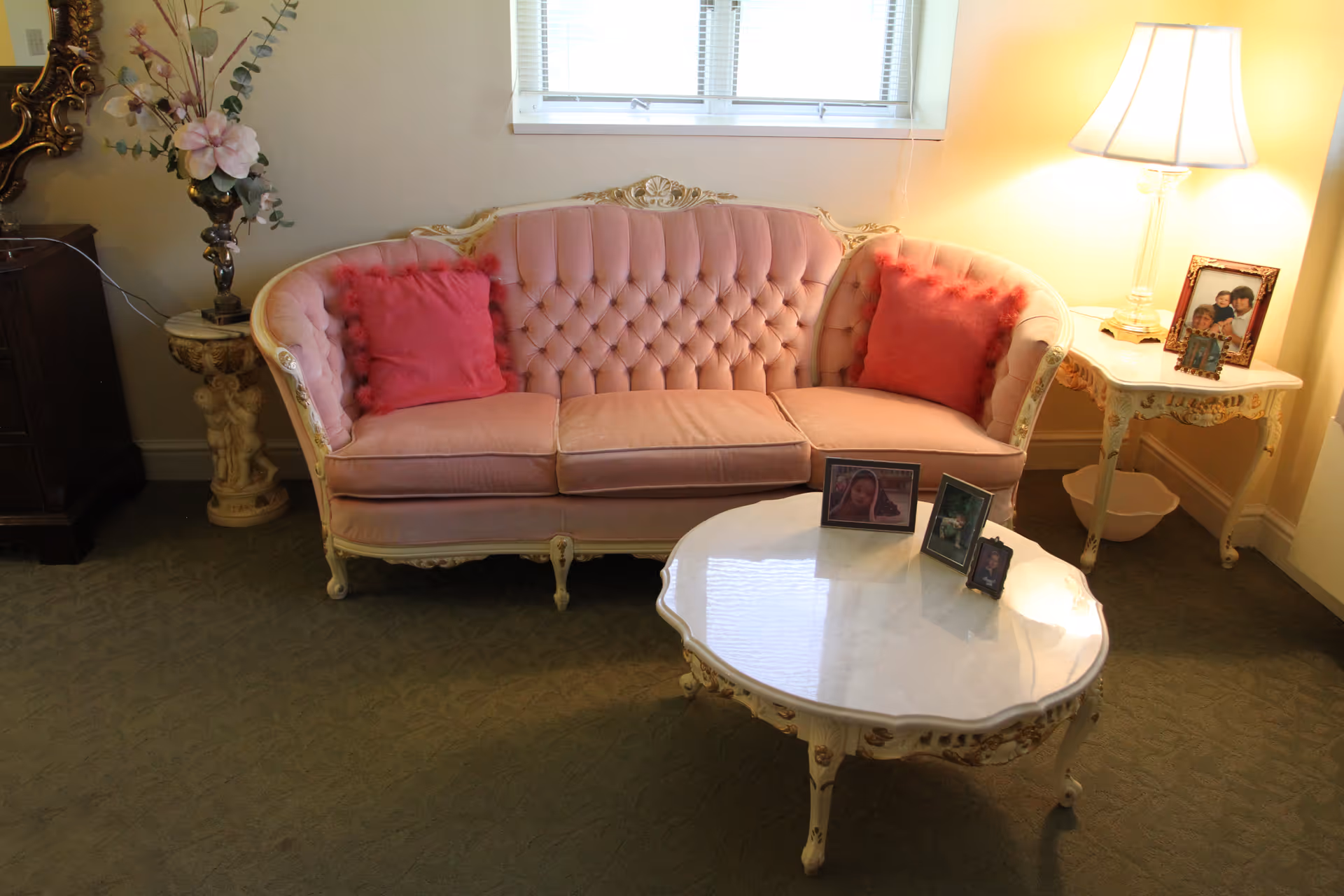 Pink tufted sofa with ornate white coffee table, lamp, and framed photos in a living room.