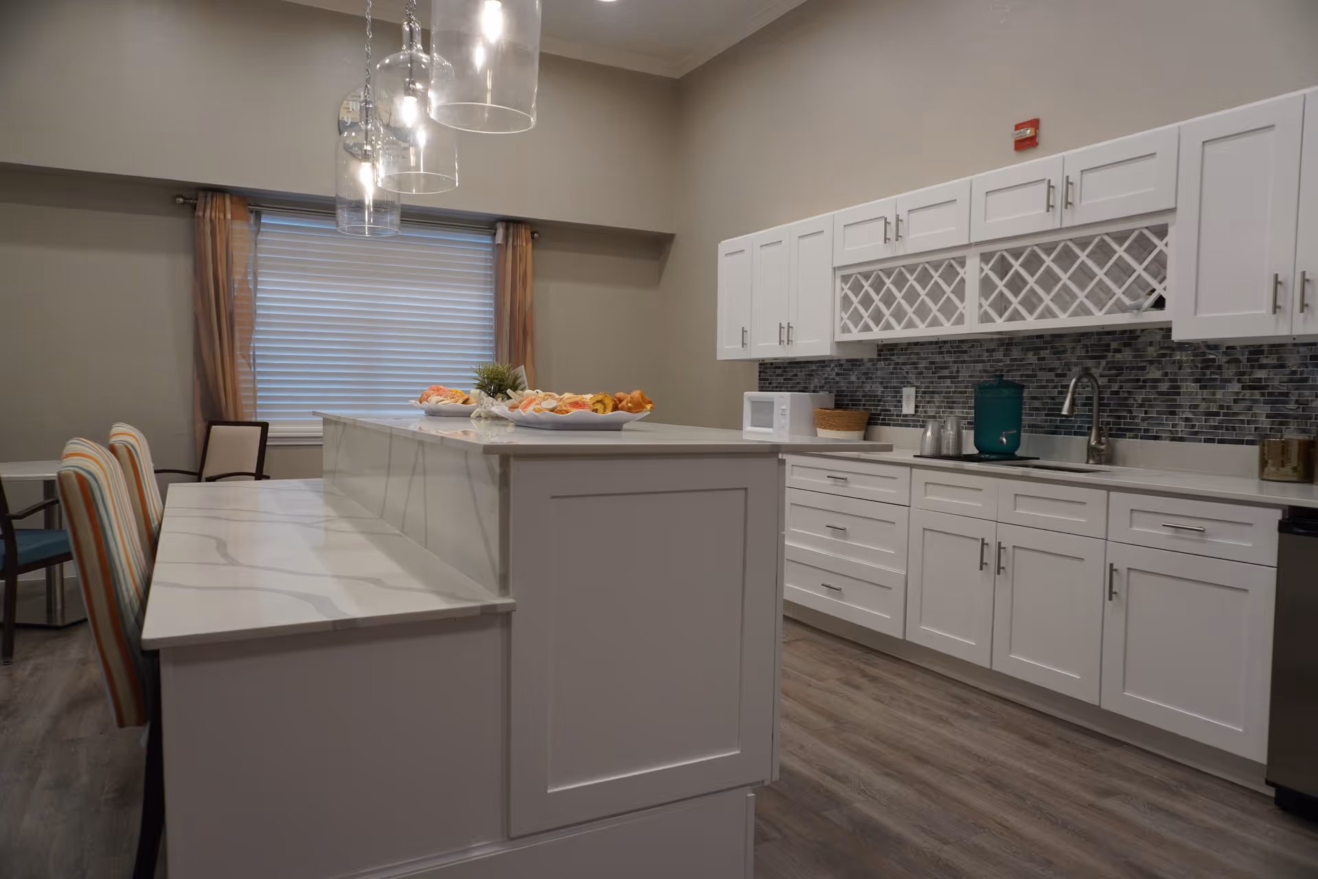 A modern kitchen area with white cabinets and drawers, a marble countertop island with bar stools, a mosaic tile backsplash, a microwave, a sink, and pendant lights hanging from the ceiling. There is a window with blinds and curtains in the background.