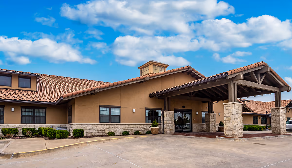 Exterior view of Chelsea Place senior living facility showing a single-story building with tan stucco walls, stone accents, a covered entrance with wooden beams, and a tiled roof under a partly cloudy blue sky.