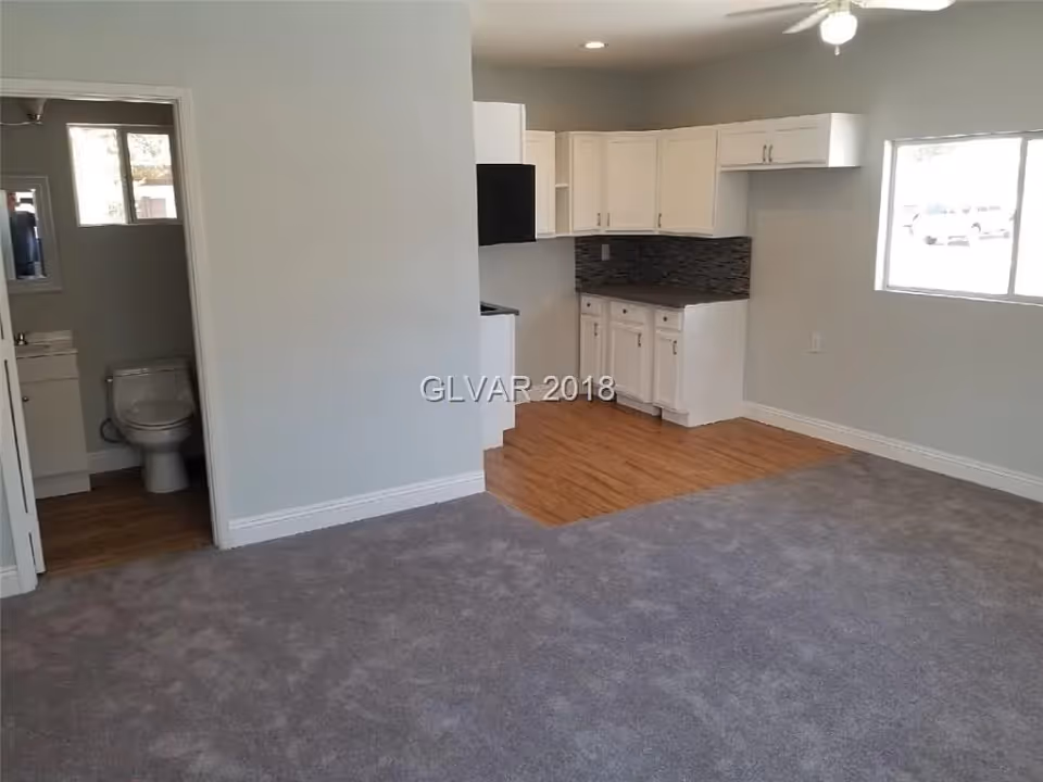 Interior view of a room with gray carpeted floor and a small kitchen area with white cabinets and a tiled backsplash. To the left, there is a bathroom with a toilet and a small window. A large window is visible on the right wall, allowing natural light into the room.