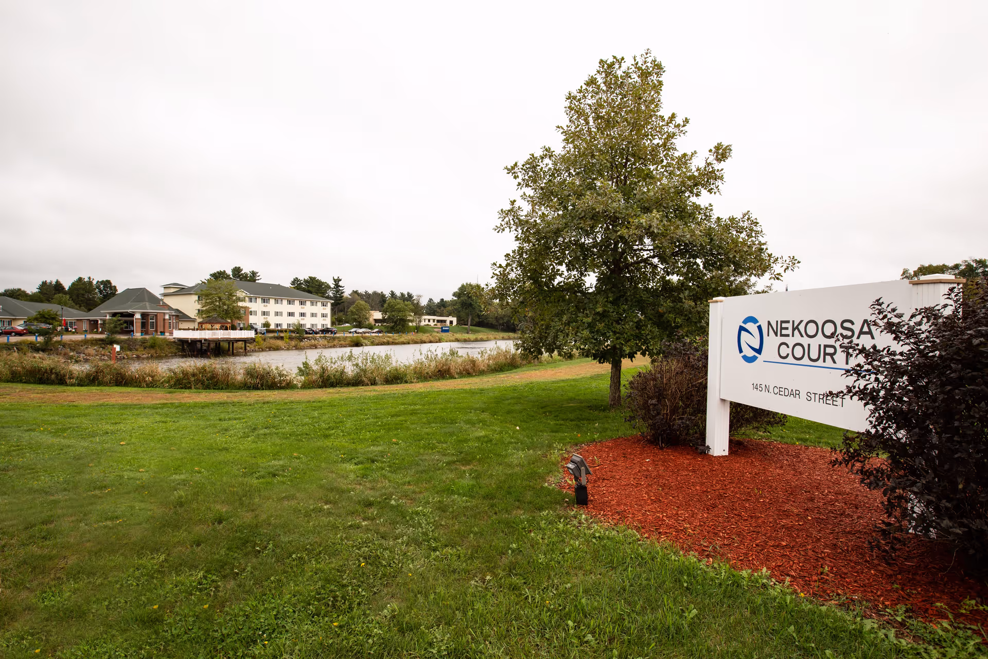 Outdoor view of Nekoosa Court facility with a large green lawn, a tree, and a white sign displaying the facility name and address. In the background, there is a pond and a multi-story building under a cloudy sky.