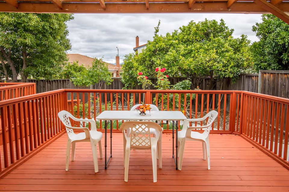 Outdoor wooden deck with a white table and four white plastic chairs. The deck is surrounded by a red wooden railing and overlooks a garden with green trees and flowering plants under a cloudy sky.