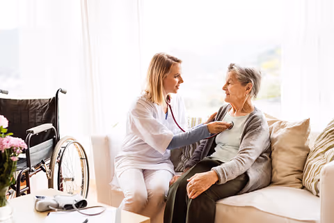 A female healthcare professional using a stethoscope to check the heartbeat of an elderly woman sitting on a couch in a bright room with a wheelchair nearby.