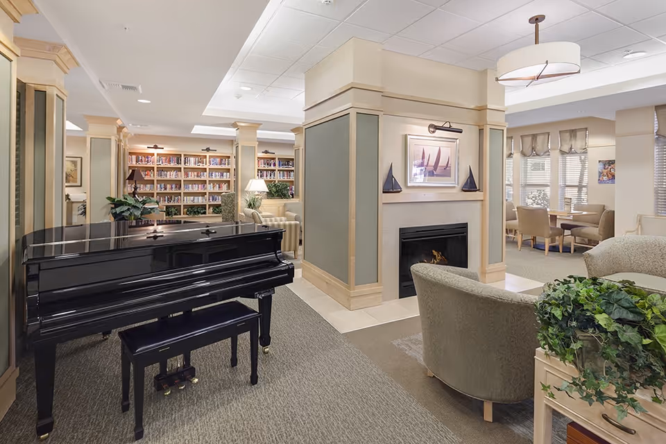 A cozy senior living common area featuring a black grand piano with a matching bench, a fireplace with a framed picture and decorative sailboats above it, comfortable armchairs, a bookshelf filled with books, and a dining area with tables and chairs near windows with blinds.