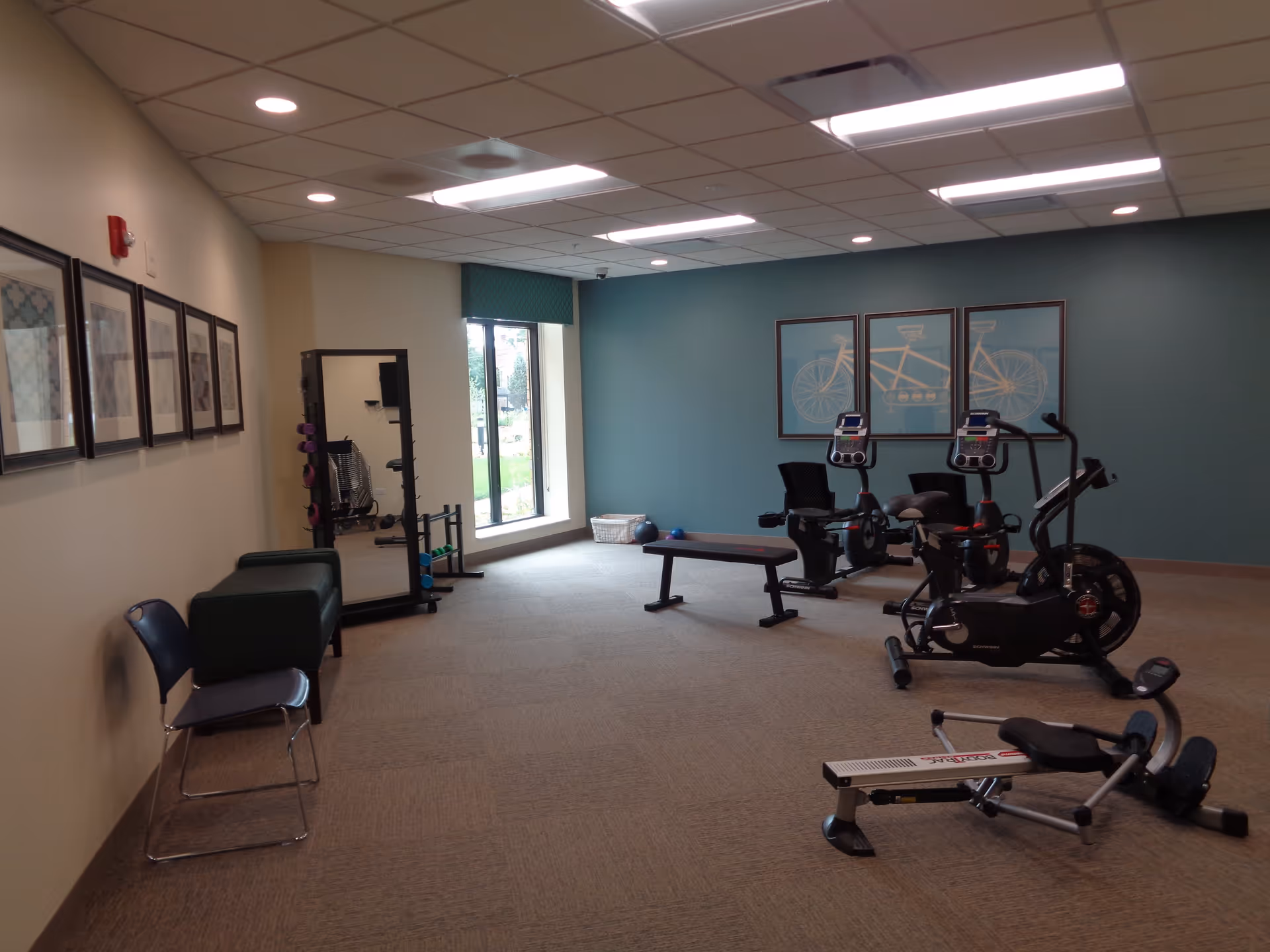 An empty fitness room with exercise bikes, a rowing machine, bench, free weights and wall art in a senior living community.