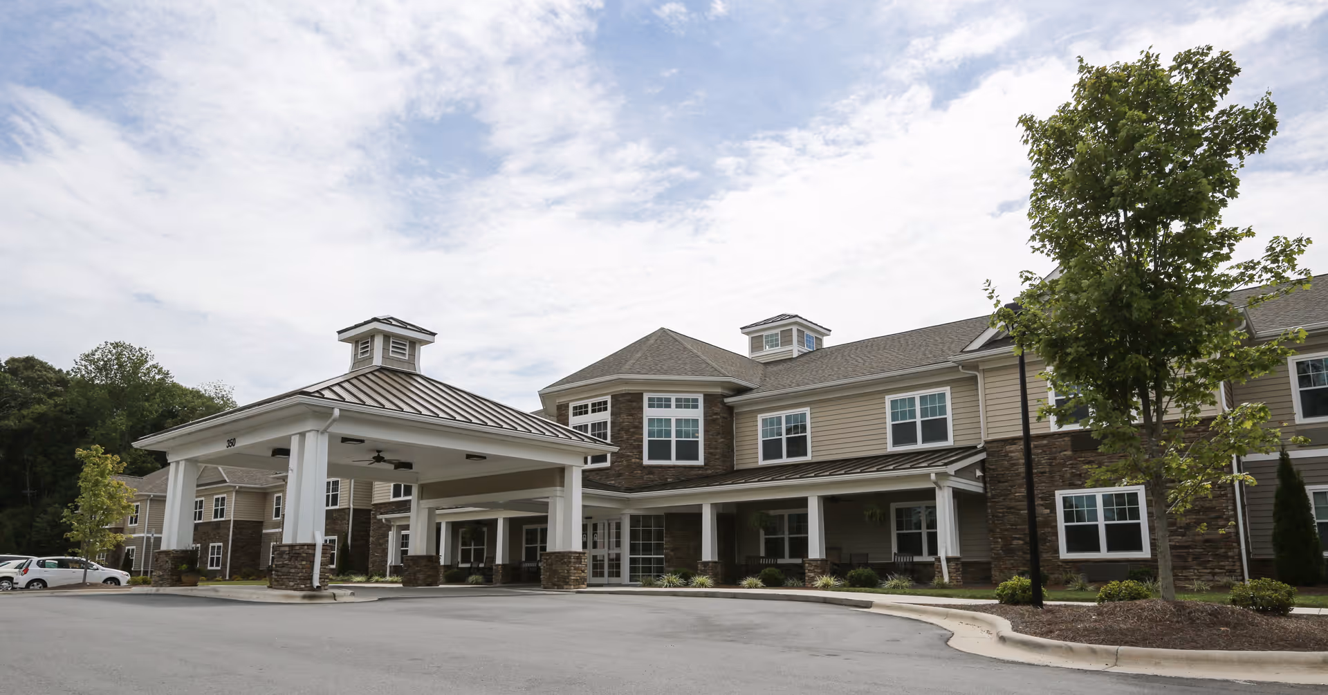 Exterior view of a two-story senior living facility building with a covered entrance, multiple windows, and a landscaped area with trees and shrubs under a partly cloudy sky.