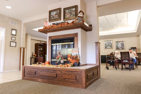 Interior view of a senior living facility featuring a wooden fireplace decorated with autumn-themed items including leaves, pumpkins, and a scarecrow doll. Two framed paintings hang above the fireplace. In the background, two elderly people are seated and conversing at a table in a common area with carpeted flooring and soft lighting.