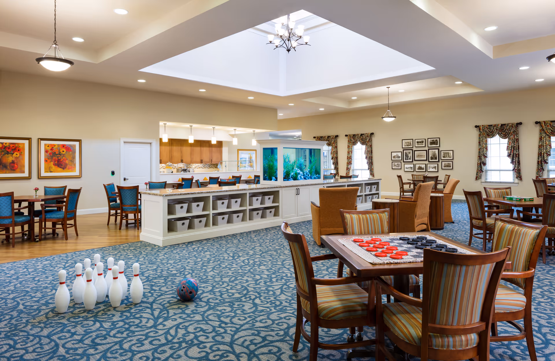 A spacious senior living community room with a patterned blue carpet and beige walls. The room features a large skylight, multiple seating areas with wooden chairs and tables, some with board games like checkers set up. There is a small bowling setup with pins and a ball on the carpet. The room also includes a large fish tank on a white cabinet, a kitchen area with bar stools, and framed artwork and photos on the walls. The space is well-lit with ceiling lights and pendant lamps.