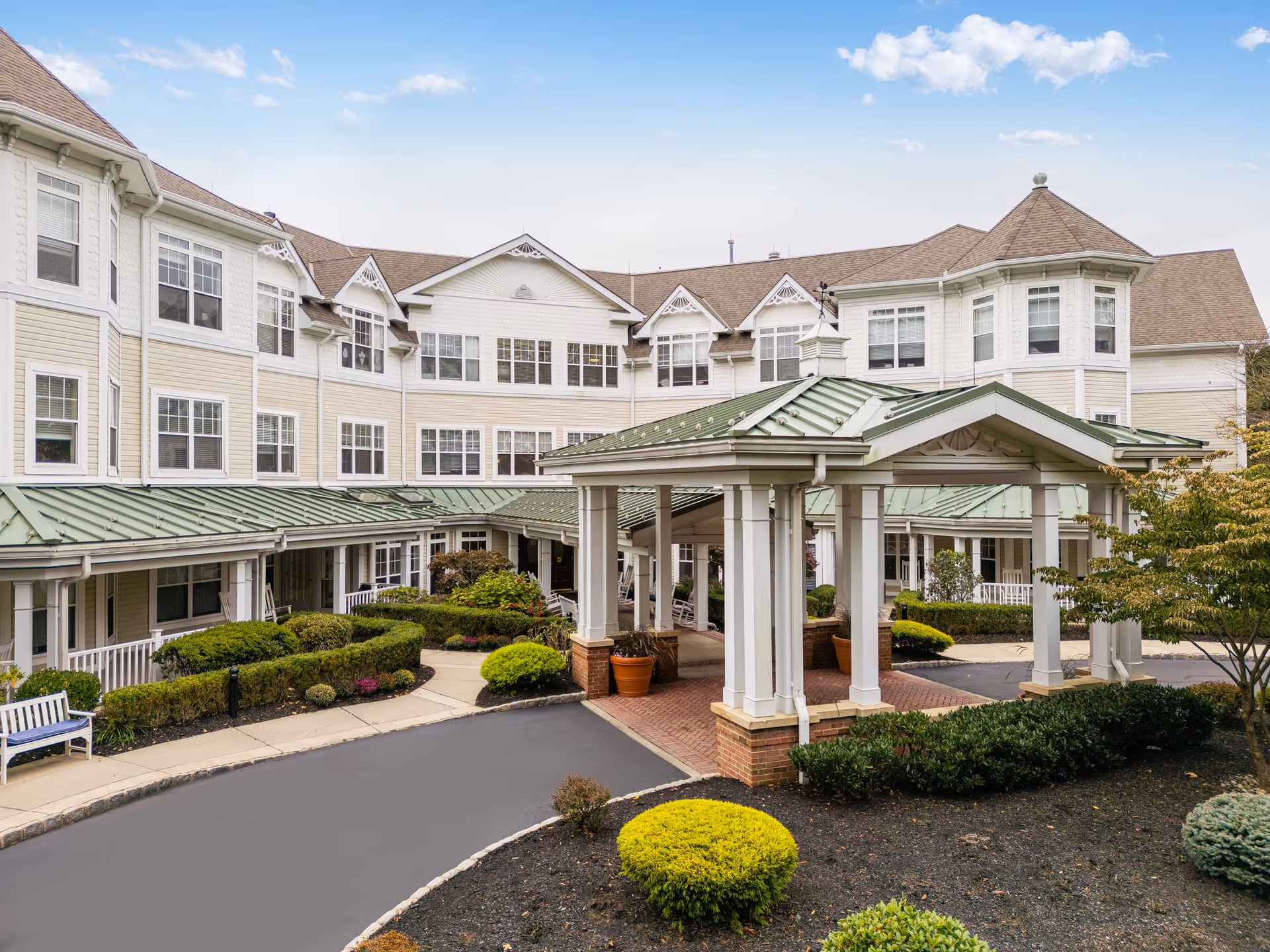 Exterior view of a senior living facility with a covered entrance, manicured bushes, and a multi-story building with numerous windows under a partly cloudy sky.