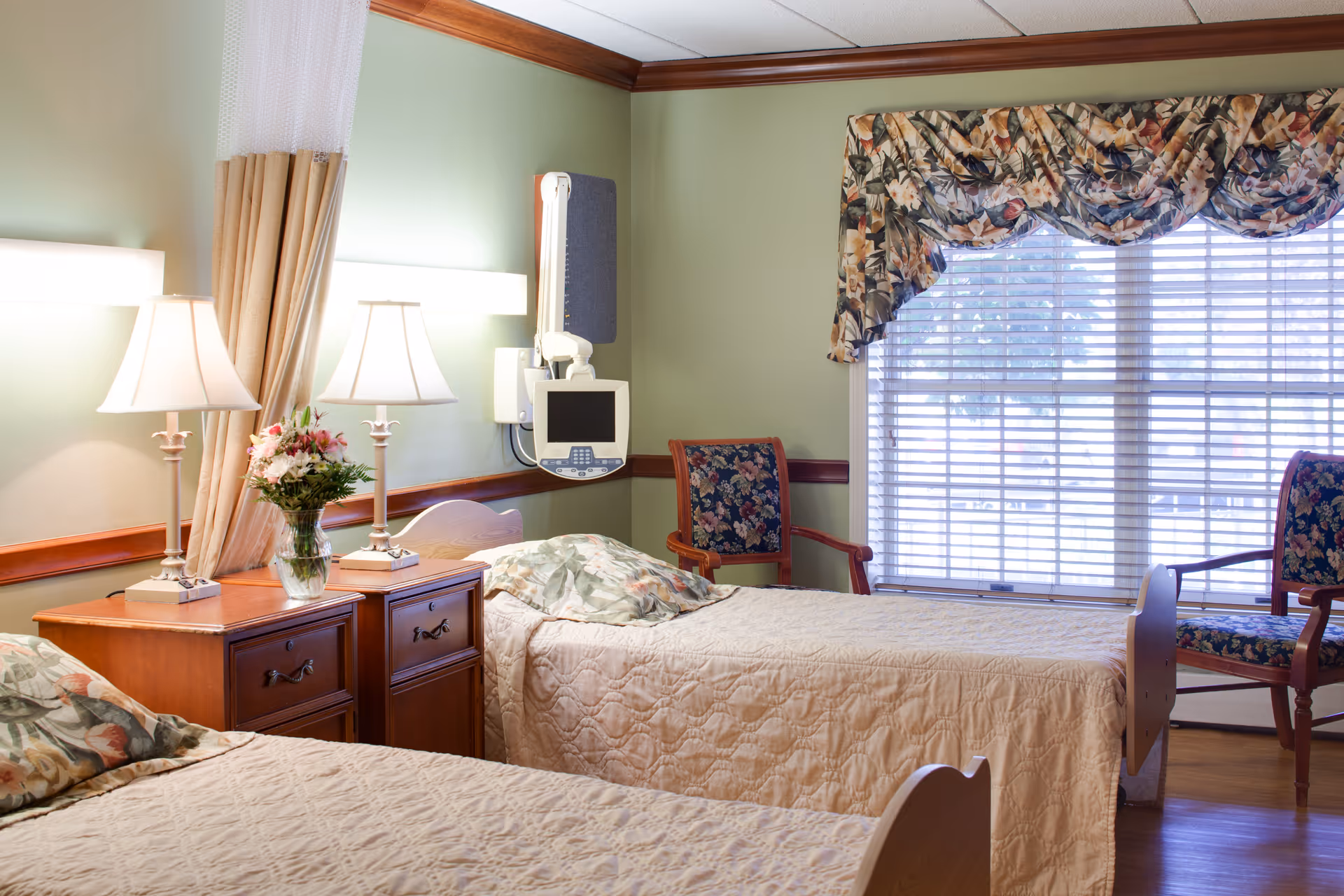 Two-bed senior living bedroom with a nightstand holding lamps and flowers, floral chairs, and a large window with blinds.