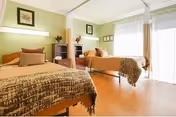Bright shared patient bedroom with two single beds, bedside tables, and large windows with sheer curtains.