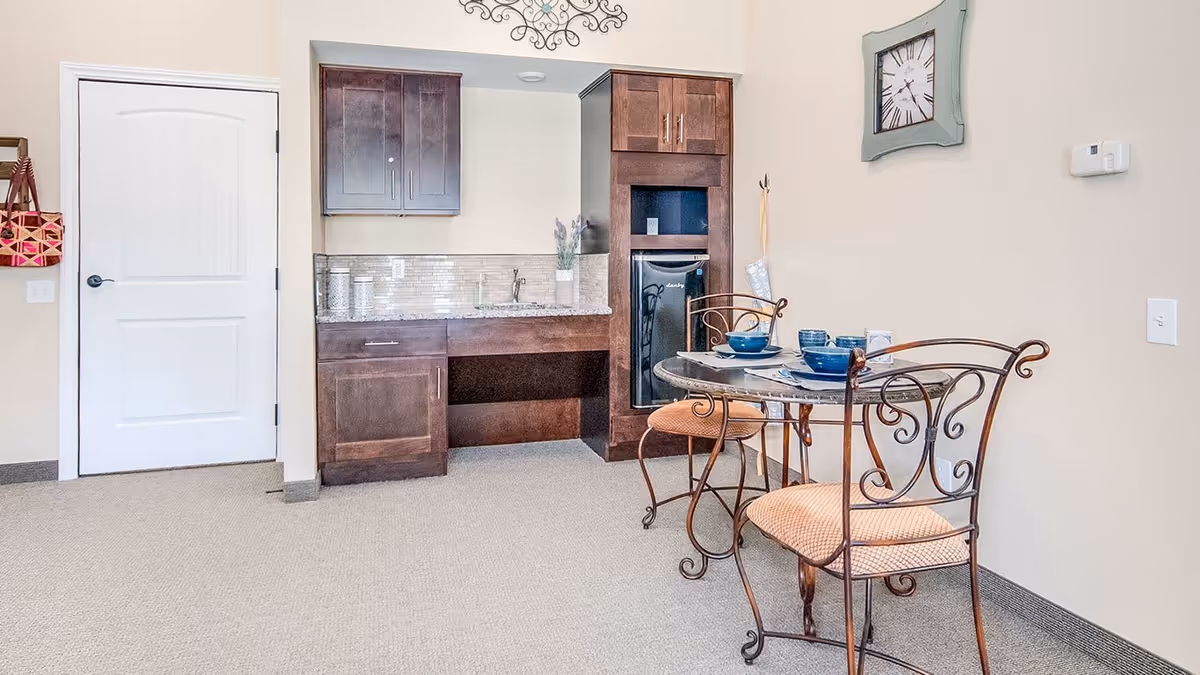 A small dining area in an assisted living facility with a round glass-top table set for two with blue bowls and plates. Two wrought iron chairs with cushioned seats are placed around the table. Behind the table is a kitchenette with dark wood cabinets, a small refrigerator, a sink, and a countertop. A decorative wall clock is mounted on the beige wall above the table. A white door and a hanging bag are visible on the left side of the image.