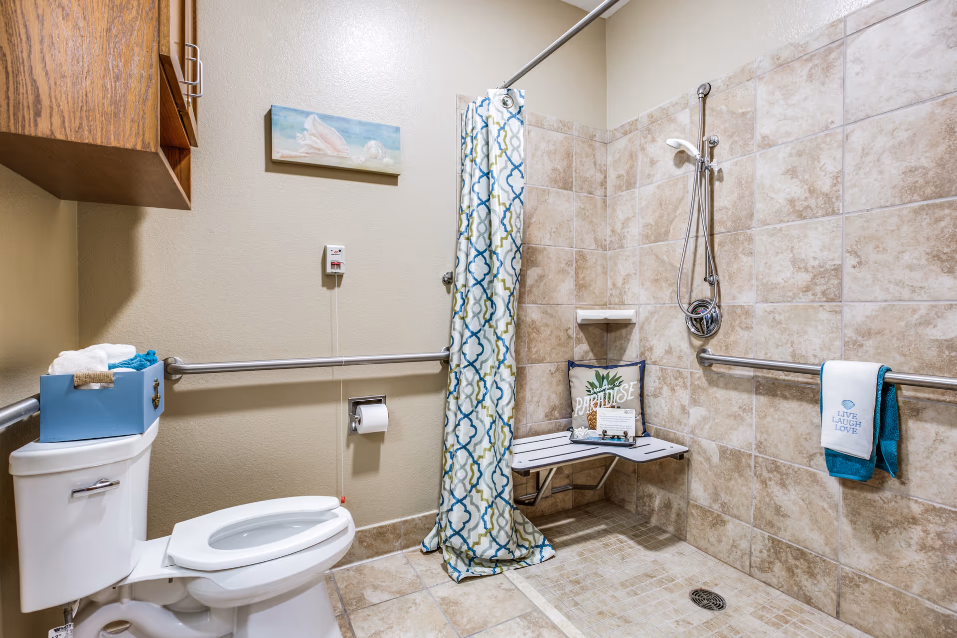 Accessible bathroom with a white toilet, grab bars on the walls, a shower area with beige tiles, a fold-down shower seat with a decorative pillow, a shower curtain with a blue and green pattern, and a towel hanging on a grab bar. There is a blue box with towels on the toilet tank and a small wall-mounted cabinet above.