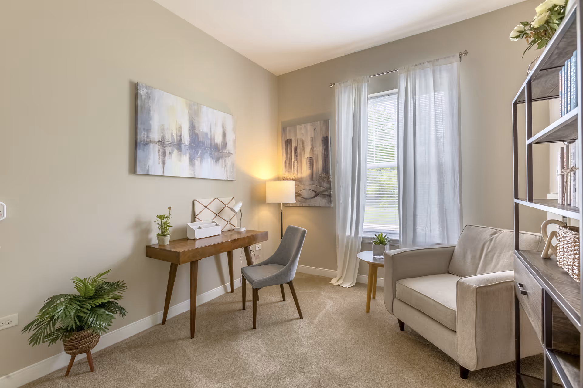 A cozy interior room with beige walls and carpet, featuring a wooden desk with a small plant, tissue box, and lamp. A gray upholstered chair is placed in front of the desk. Next to a window with sheer white curtains, there is a small round table with a plant and a beige armchair. A tall bookshelf with books and decorative items is on the right side of the image. Two abstract paintings hang on the walls, and a floor lamp provides warm lighting.