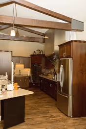 Open kitchen with dark wood cabinets, stainless-steel refrigerator, island countertop, and exposed ceiling beams.