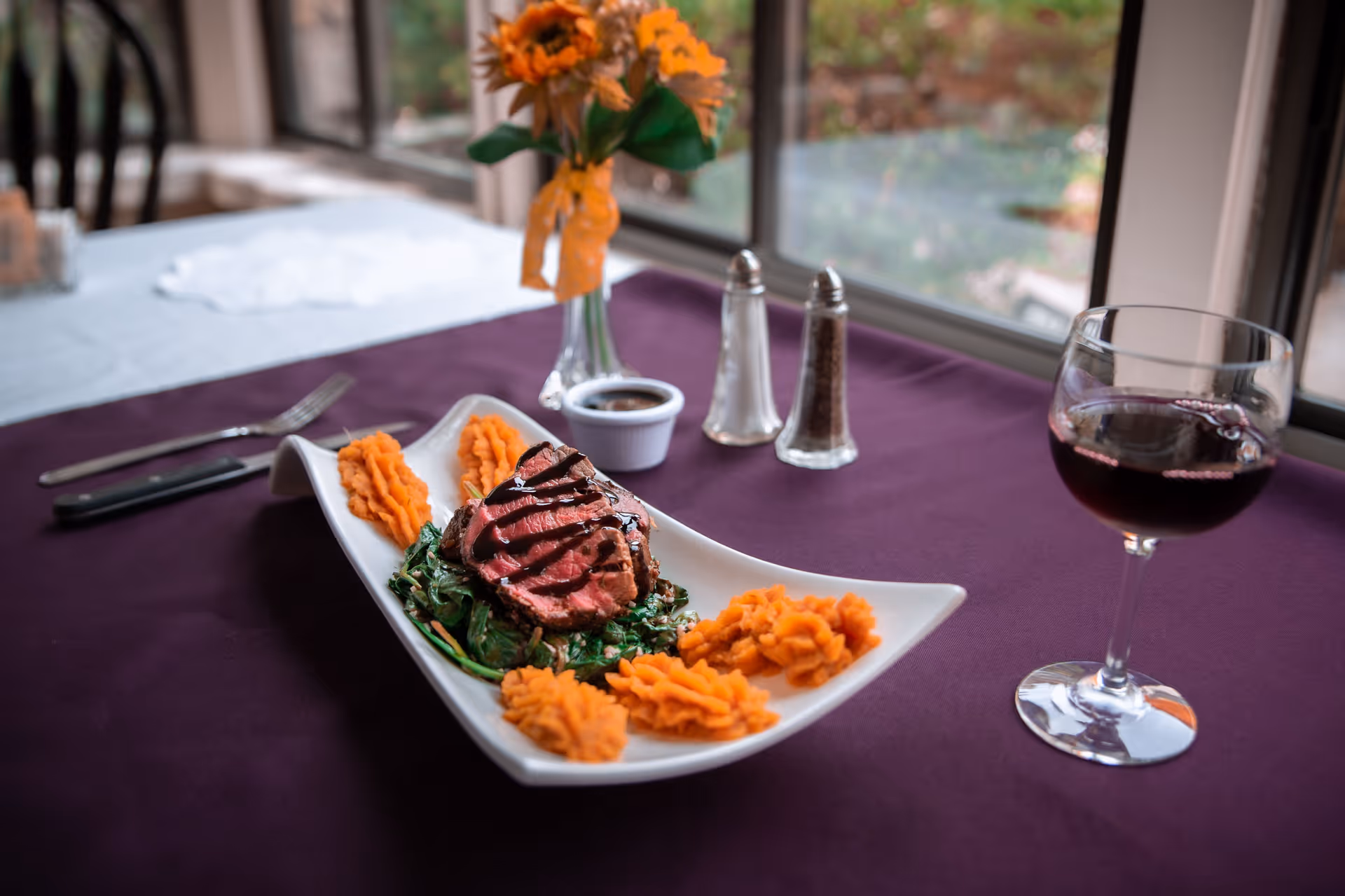 Plated sliced steak on greens with mashed sweet potatoes, a glass of red wine and condiments on a purple-tablecloth dining table by a window.
