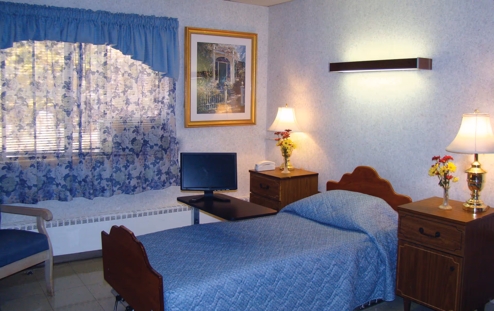 Single-occupancy bedroom with a blue bedspread, wooden headboard and nightstands, lamps, small TV, and floral curtains.