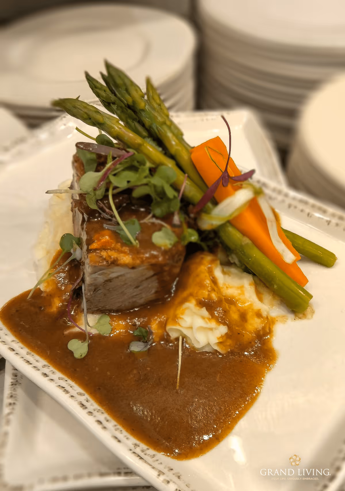 A plated meal featuring a piece of meat with brown gravy, mashed potatoes, steamed asparagus, and carrots garnished with microgreens on a white square plate. Stacks of white plates are visible in the background.