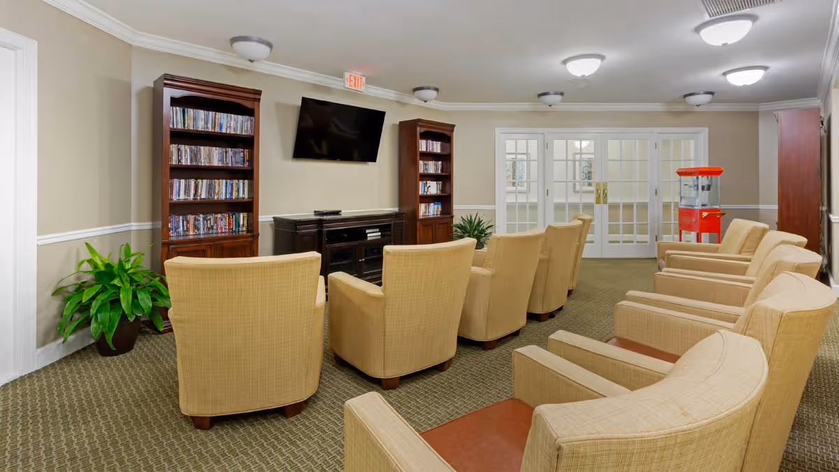 A senior living common room with rows of beige armchairs facing a wall-mounted TV, bookshelves, and a popcorn machine near glass doors.