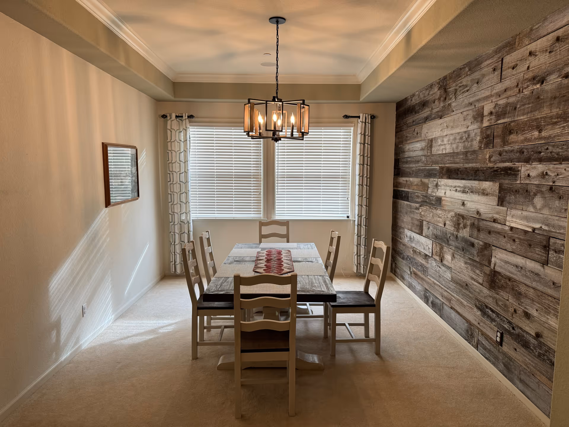 A dining room with a rectangular wooden table and six chairs. The table has a red and white patterned runner. There is a modern chandelier hanging above the table. The room features a large window with white blinds and patterned curtains. One wall is decorated with rustic wooden planks, while the other walls are painted beige. A framed mirror hangs on one wall.