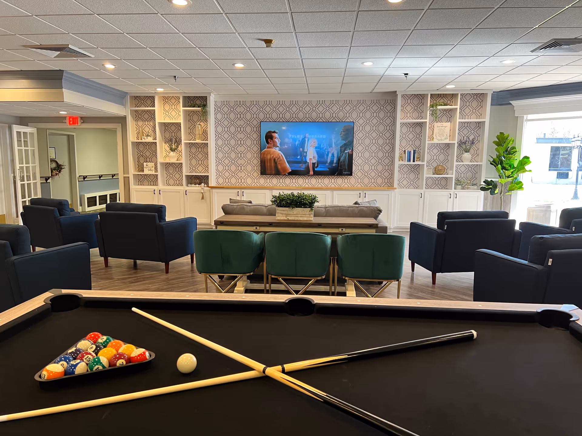 Communal lounge with a pool table in the foreground, sofas and chairs facing a wall-mounted TV and decorative shelving.
