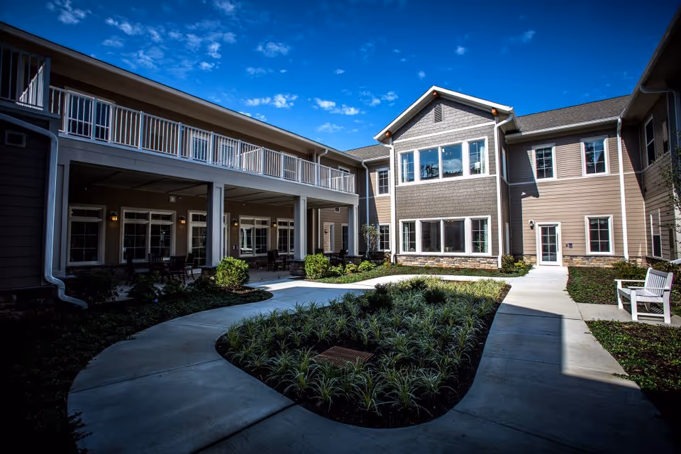 Outdoor courtyard area of a senior living facility with a curved concrete walkway, landscaped plants, benches, and a two-story building with multiple windows and a balcony under a blue sky with some clouds.