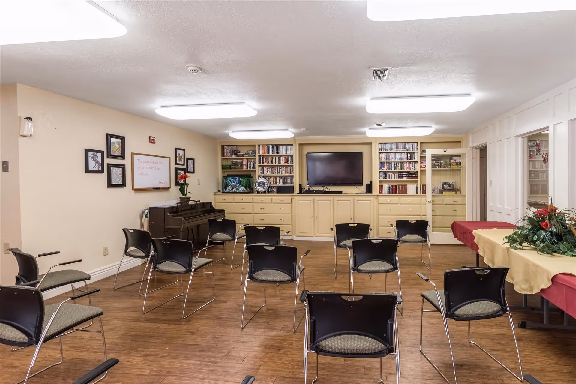 A community room with multiple black chairs arranged in rows facing a wall with built-in cabinets, bookshelves, a flat-screen TV, and a fish tank. There is a piano against the left wall with framed pictures and a whiteboard above it. On the right side, there are tables covered with red and yellow tablecloths and a floral arrangement. The room has wood flooring and bright ceiling lights.