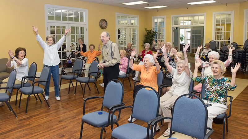 A group of elderly people participating in a seated exercise or activity session in a bright room with wooden floors and multiple chairs. Some participants are seated while others stand, all raising their arms enthusiastically. The room has large windows and glass-paneled doors in the background.
