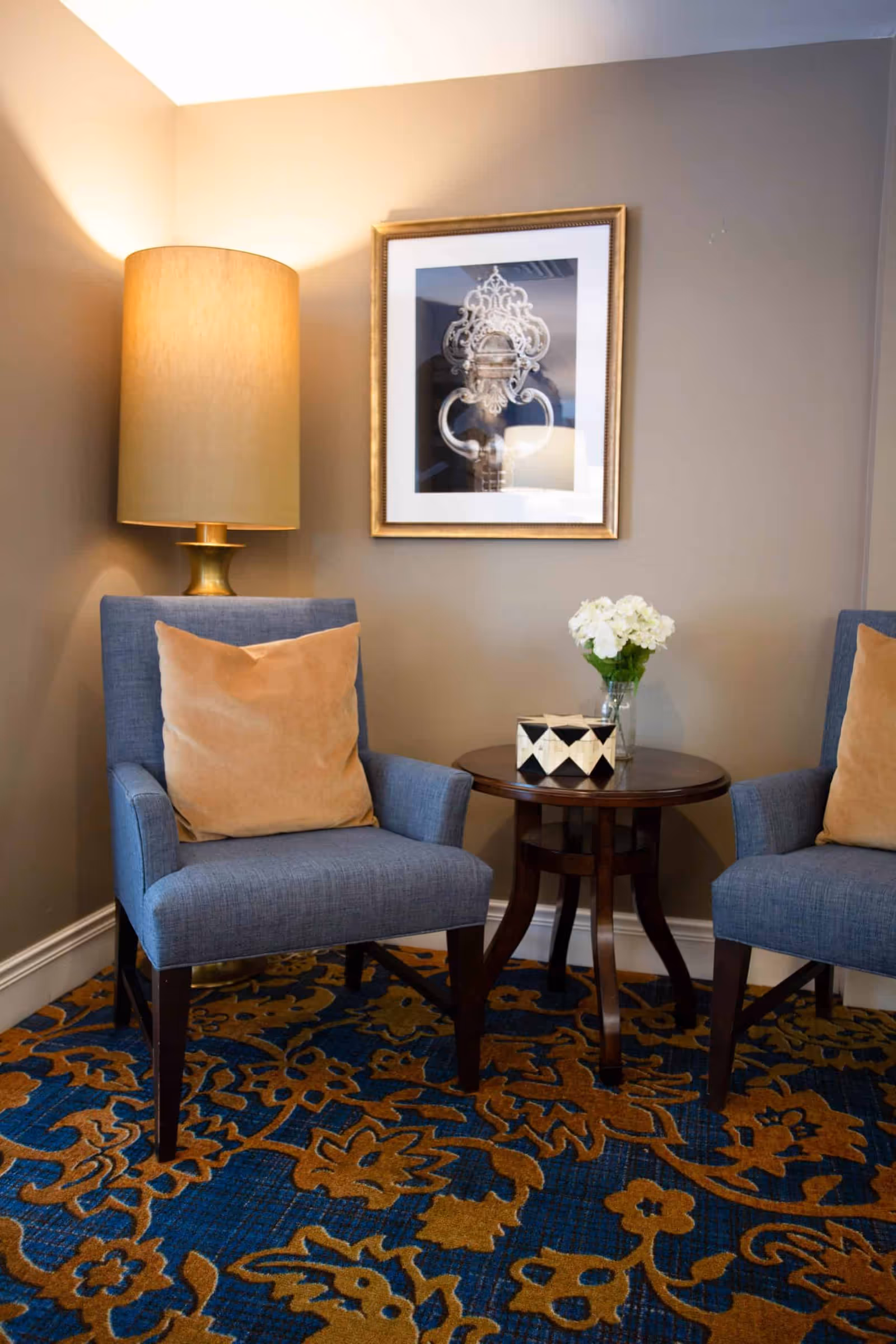 A cozy corner with two blue upholstered armchairs each with a beige pillow, a round wooden side table between them holding a small decorative box and a vase with white flowers, a tall beige lampshade on a brass lamp behind one chair, and a framed artwork on the beige wall above.