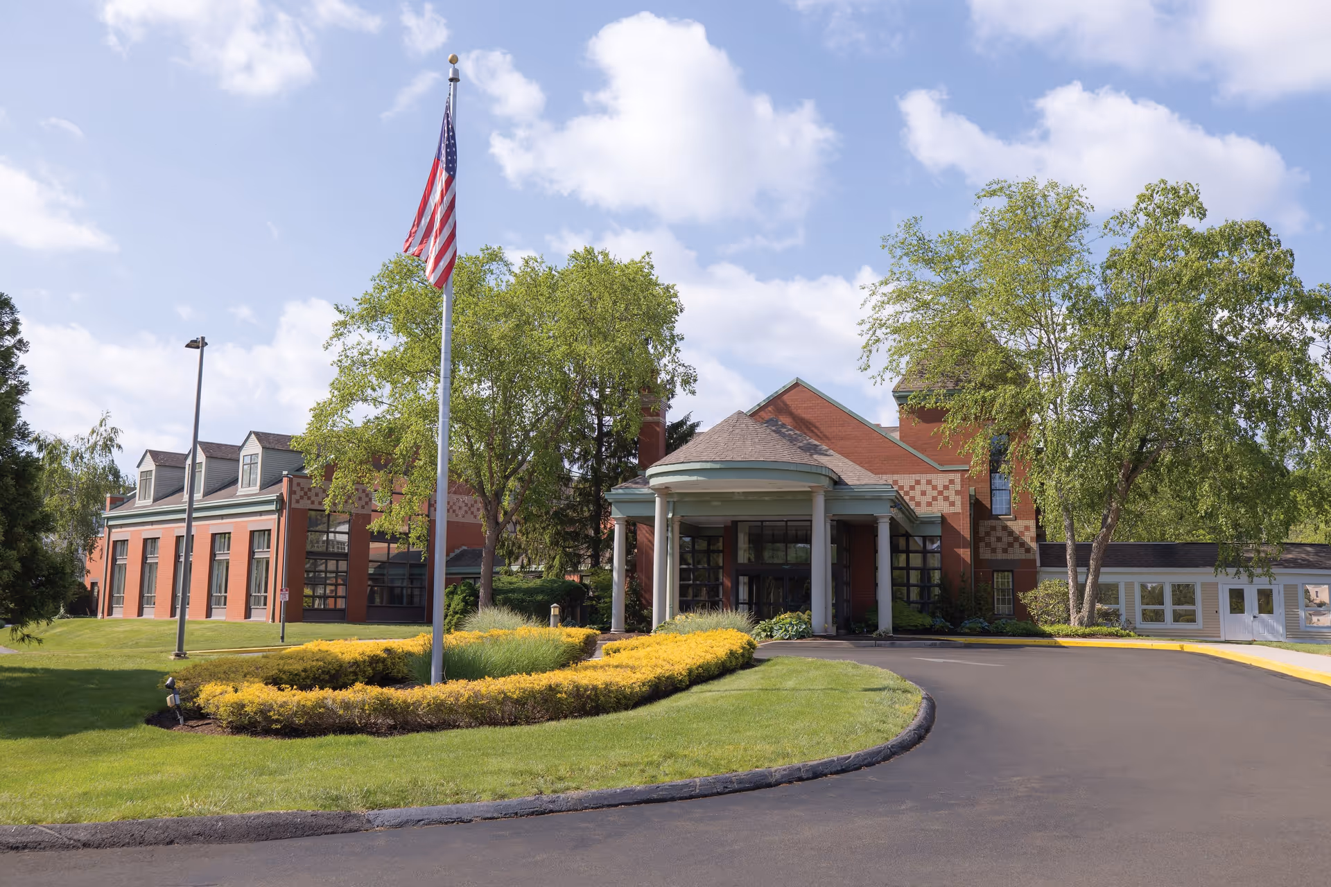 Front exterior view of a brick building with a covered entrance supported by white columns, surrounded by green trees and well-maintained landscaping including a circular flower bed with yellow bushes. An American flag is flying on a flagpole in front of the building under a partly cloudy sky.