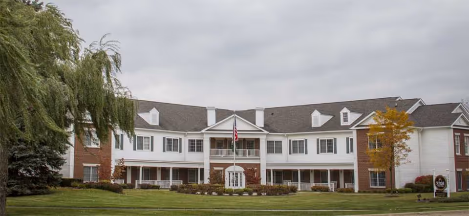 Exterior view of a large two-story senior living facility building with white siding and brick accents, a central entrance with columns, an American flag on a flagpole in front, surrounded by green lawn and trees under a cloudy sky.
