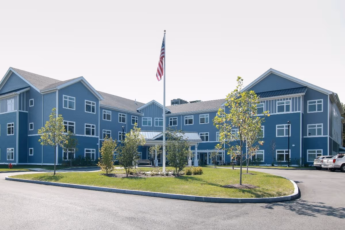 Exterior view of a three-story senior living facility building painted in blue with white trim. The building surrounds a circular driveway with a grassy island in the center featuring small trees and an American flag on a flagpole. Several cars are parked on the right side of the building.