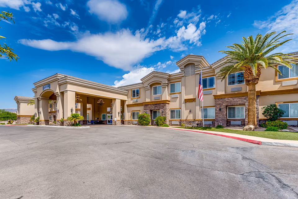 Front exterior of San Martin Senior Living showing the entrance porte-cochère, American flag, and a palm tree under a bright blue sky.