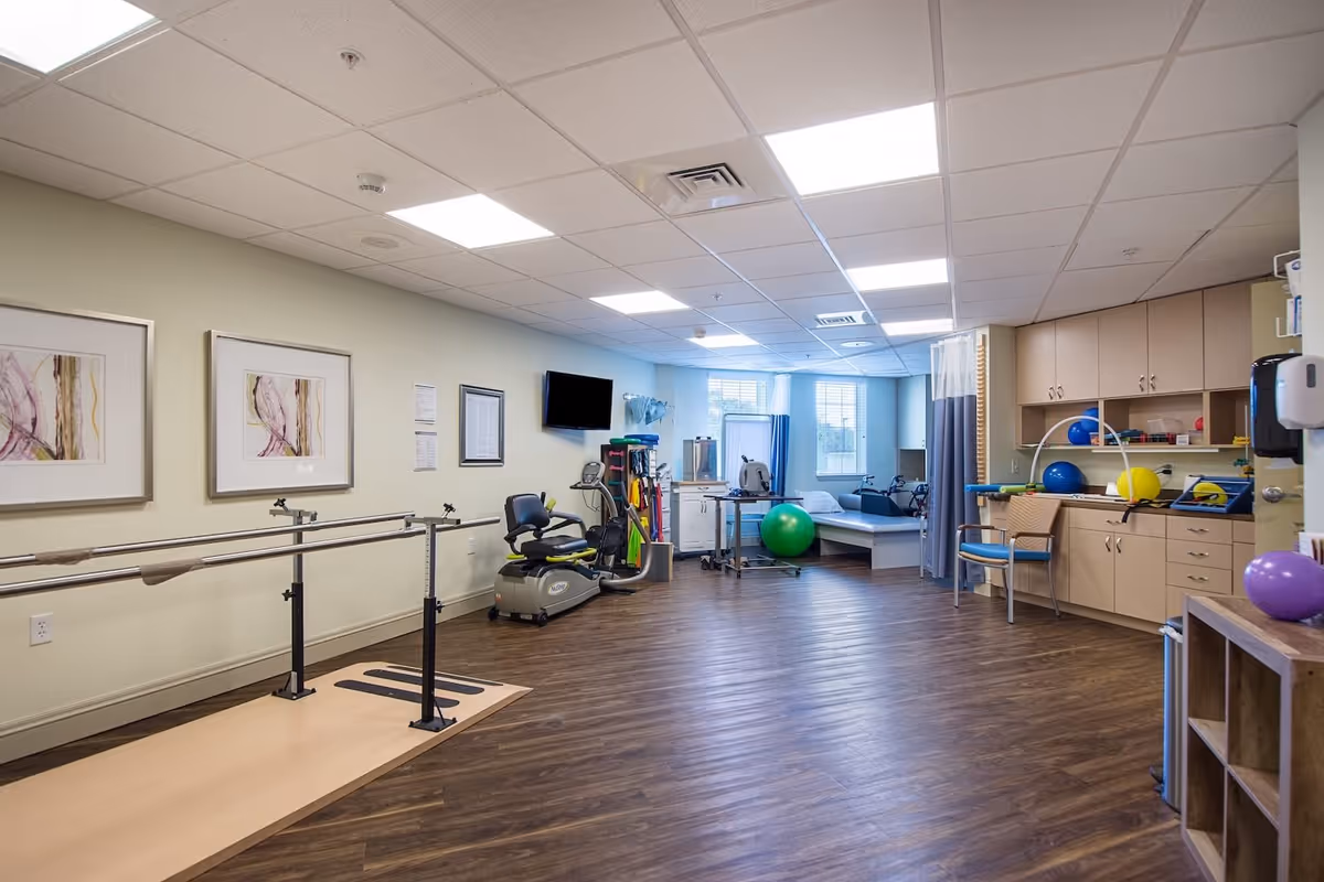 A spacious physical therapy or rehabilitation room with wooden flooring, parallel bars for walking exercises, an exercise bike, various therapy balls, and medical equipment. The room has beige walls with framed artwork, a wall-mounted TV, and large windows letting in natural light. Cabinets and a chair are visible on the right side.