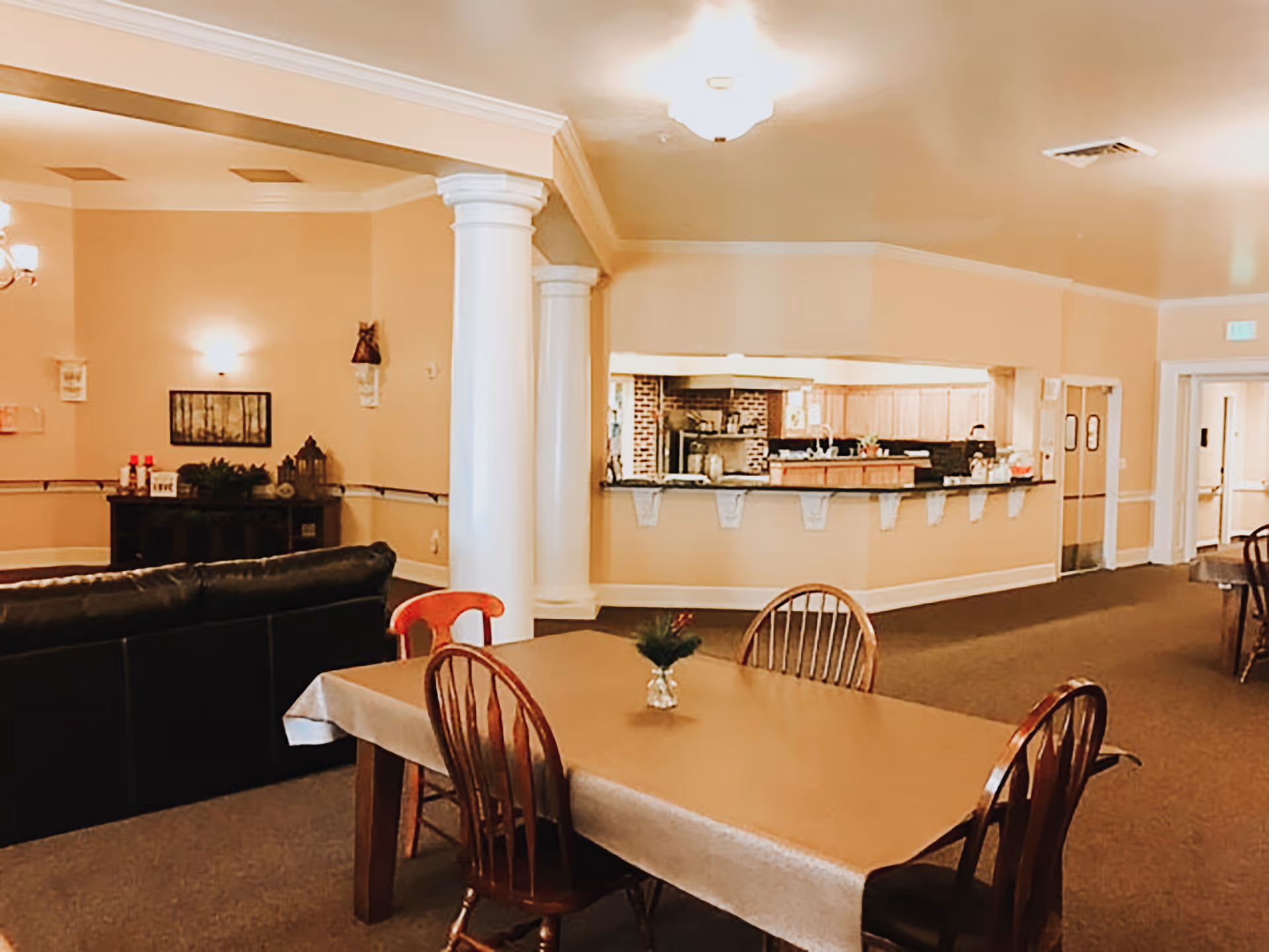 Interior view of a common area in an assisted living facility featuring a dining table with four wooden chairs and a small centerpiece. In the background, there is a kitchen area with a counter and cabinets, as well as a black leather couch and decorative columns. The walls are painted beige, and the room is warmly lit with ceiling lights and wall sconces.