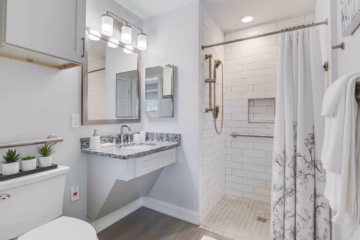 A modern bathroom featuring a white toilet with small potted plants on top, a granite countertop with a sink and soap dispenser, a large mirror with three light fixtures above it, and a walk-in shower with white subway tiles, a handheld showerhead, grab bars, and a white shower curtain with a floral pattern.