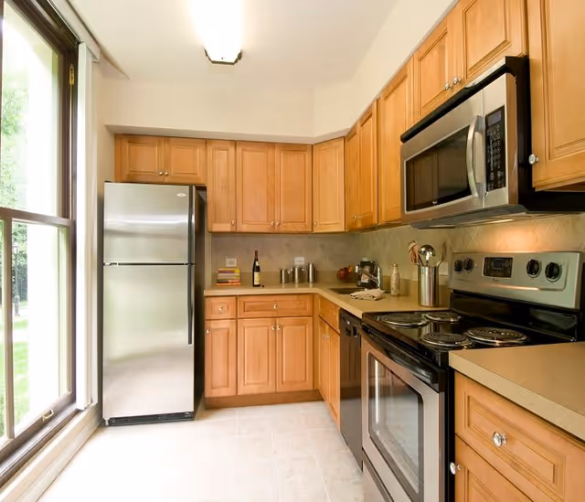 A modern kitchen with wooden cabinets, a stainless steel refrigerator, microwave, and electric stove. There is a large window on the left side letting in natural light, and various kitchen utensils and items are placed on the countertops.