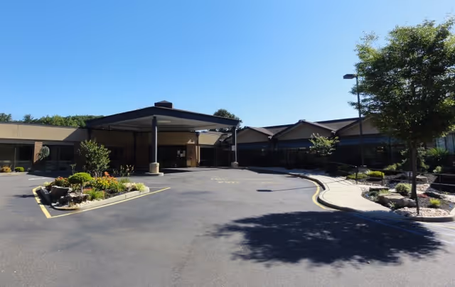 Front exterior view of a single-story building with a covered entrance, surrounded by landscaped greenery and a paved driveway under a clear blue sky.