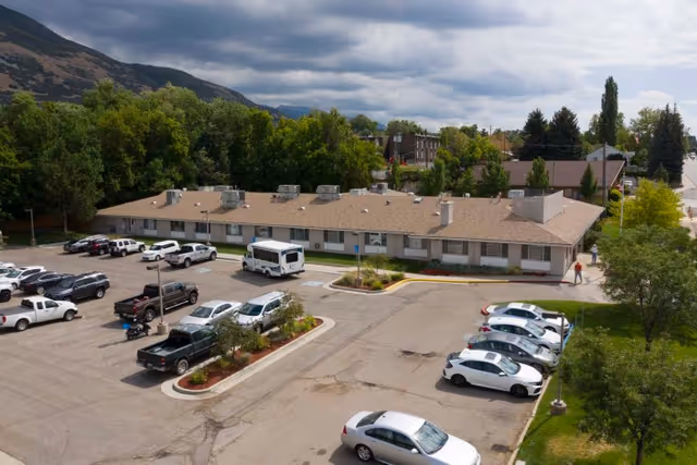 Aerial view of a single-story healthcare and rehabilitation facility building with a beige roof, surrounded by parked cars in a parking lot. The building is set against a backdrop of trees and hills under a cloudy sky.