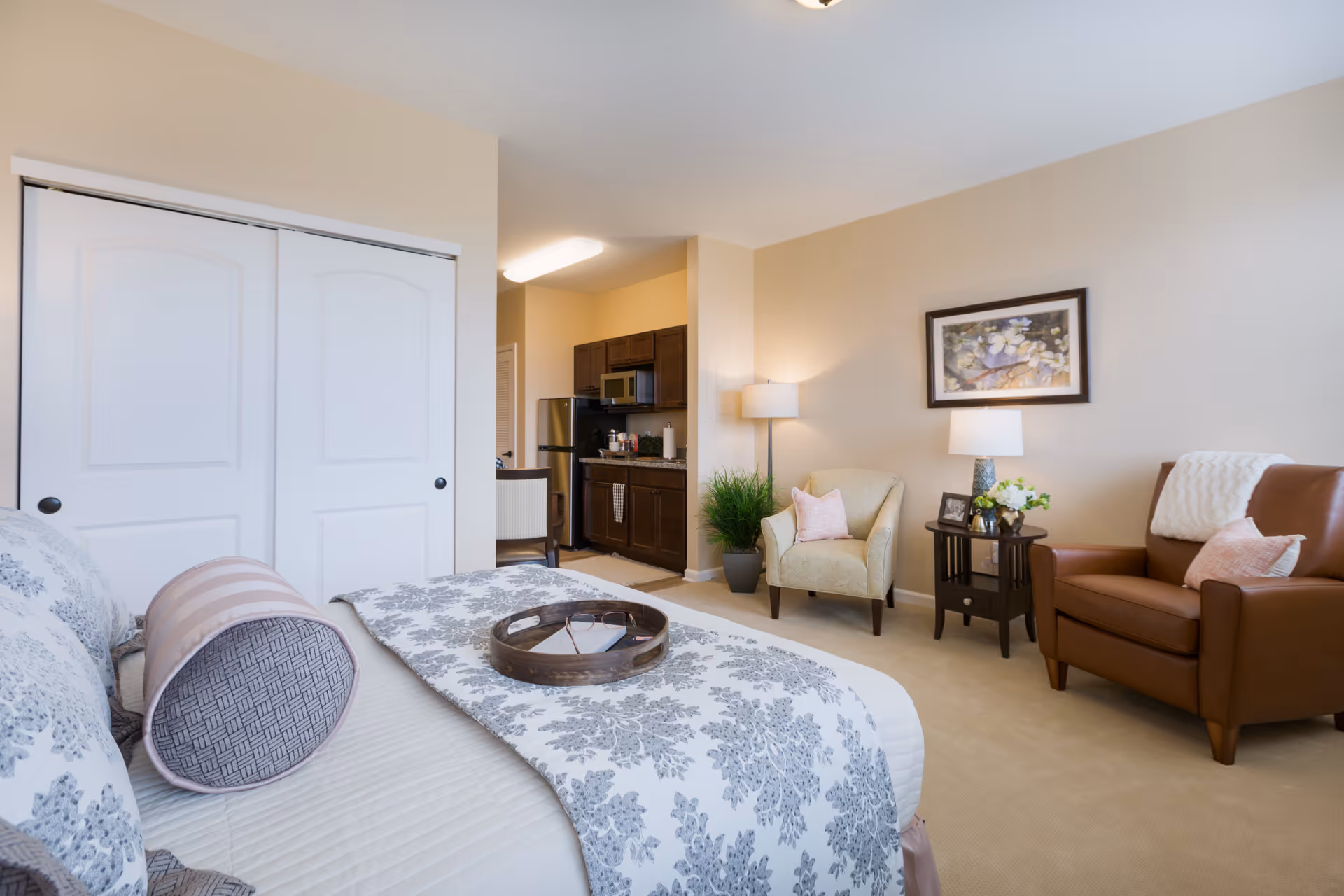 A cozy senior living studio apartment featuring a bed with patterned bedding and a decorative bolster pillow, a small kitchenette with dark cabinets and stainless steel appliances, a beige armchair and a brown leather armchair with pink pillows, a side table with a lamp and flowers, a floor lamp, a framed floral painting on the wall, and a closet with white sliding doors.