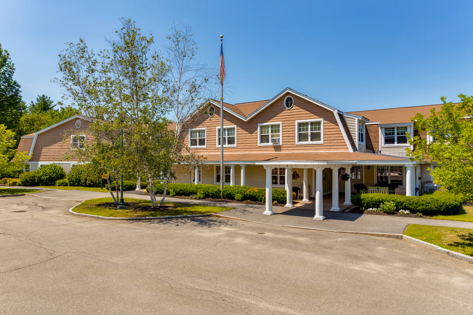 Exterior view of Chase Point Assisted Living facility showing a two-story building with beige siding and white trim, a covered entrance supported by white columns, an American flag on a flagpole, surrounded by trees and greenery under a clear blue sky.