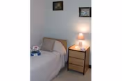 A small bedroom with a single bed covered in a light-colored bedspread. Next to the bed is a wooden nightstand with three drawers, topped with a lit table lamp and a small decorative item. Two framed pictures hang on the light-colored wall above the bed.