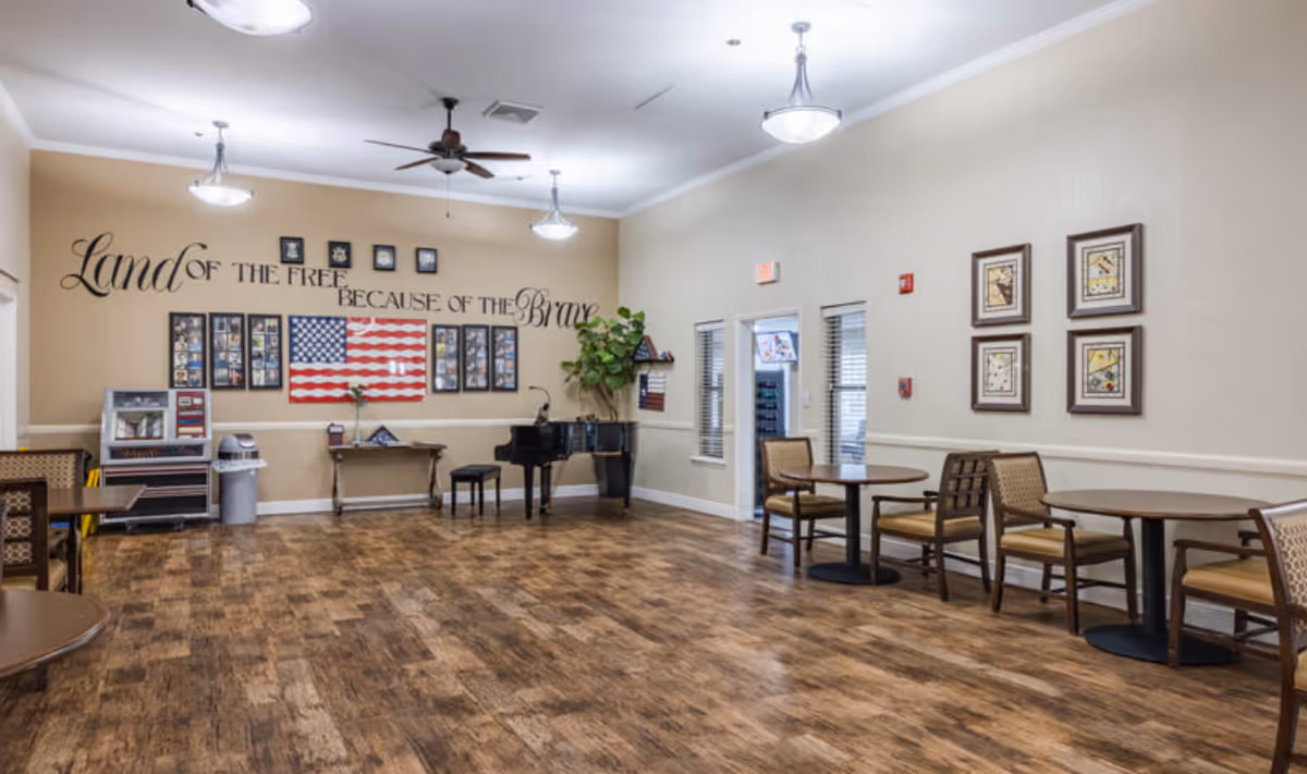 A spacious room with wooden flooring and beige walls featuring a patriotic wall decal that reads 'Land of the Free Because of the Brave' above framed photos and an American flag. The room contains several round tables with chairs, a black grand piano with a stool, a small table with a plant, and ceiling fans with lights.