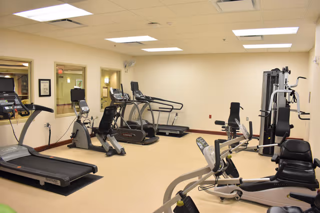 A fitness room with various exercise equipment including treadmills, stationary bikes, and weight machines. The room has beige walls, a beige floor, and fluorescent ceiling lights. There are windows on one wall looking into adjacent rooms.