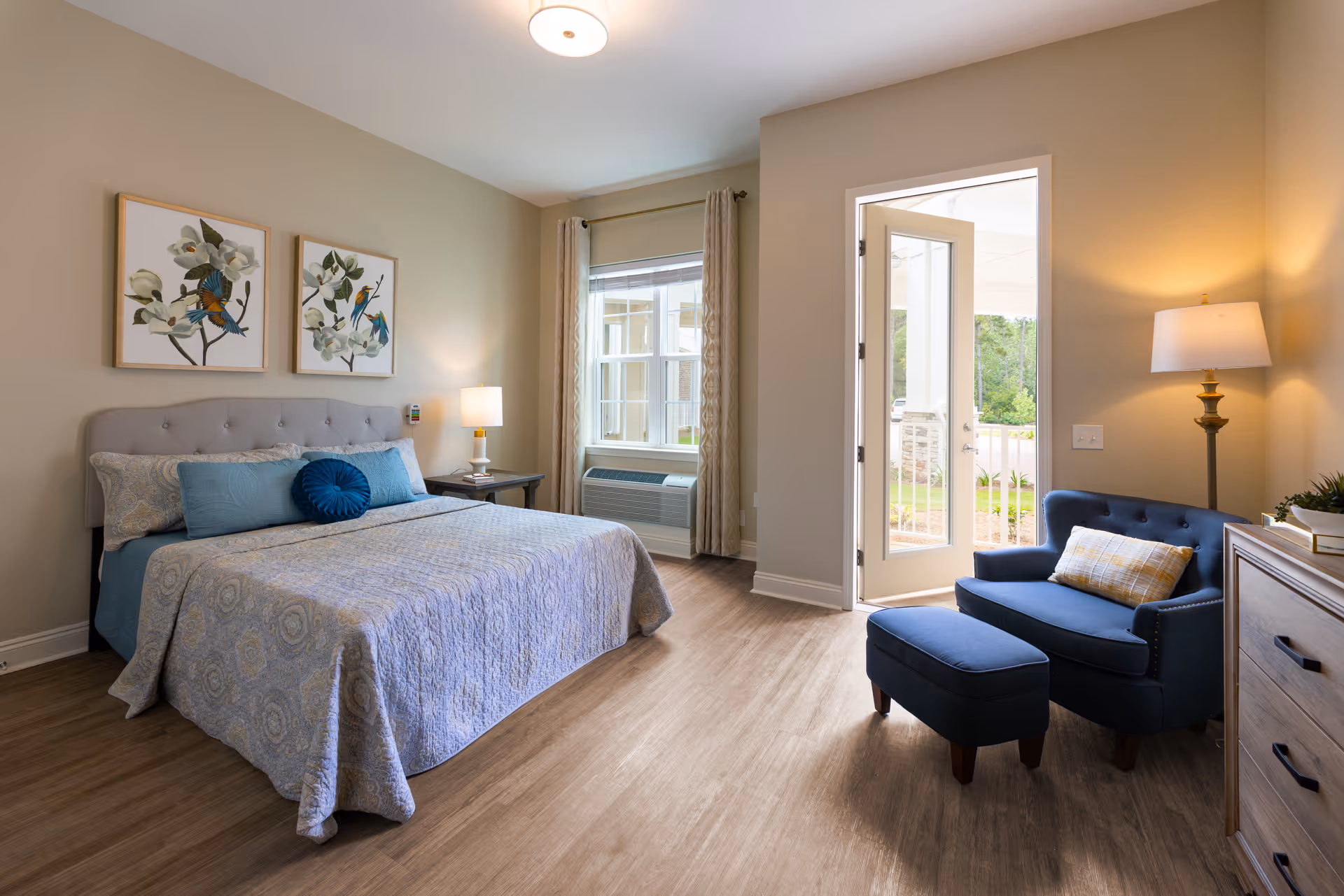 A bright and cozy bedroom with a large bed covered in a patterned quilt and multiple pillows. Above the bed are two framed artworks featuring birds and flowers. To the right of the bed is a window with curtains and an air conditioning unit below it. Next to the window is a glass door leading outside. In the corner near the door is a blue upholstered armchair with a matching ottoman and a plaid pillow, accompanied by a floor lamp. A wooden dresser is partially visible on the right side of the image. The room has wood flooring and neutral-colored walls.