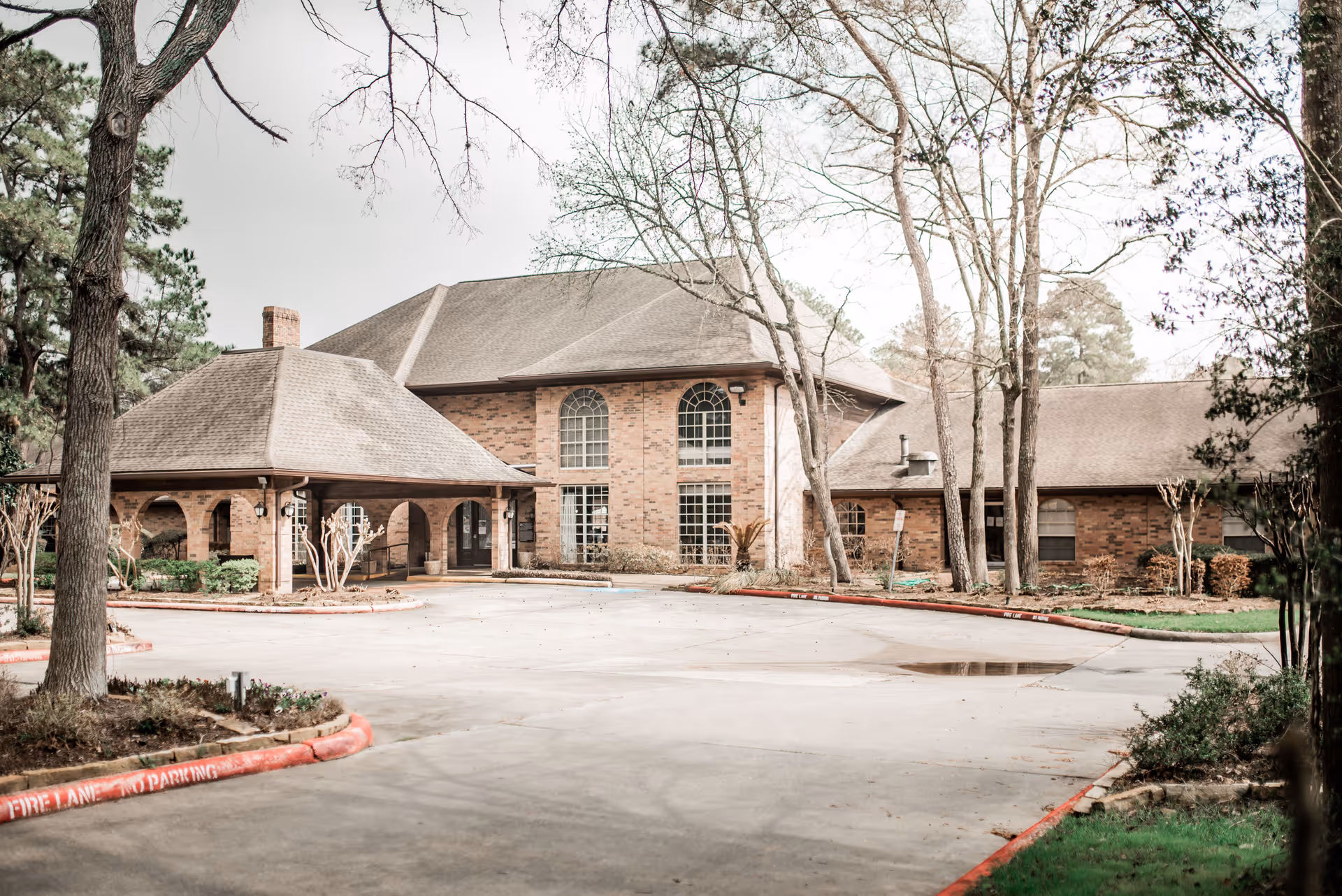 Exterior view of The Woodlands Nursing and Rehabilitation Center showing a large brick building with multiple windows and a covered entrance. The surrounding area includes trees, shrubs, and a paved driveway with a red curb marked as a fire lane.