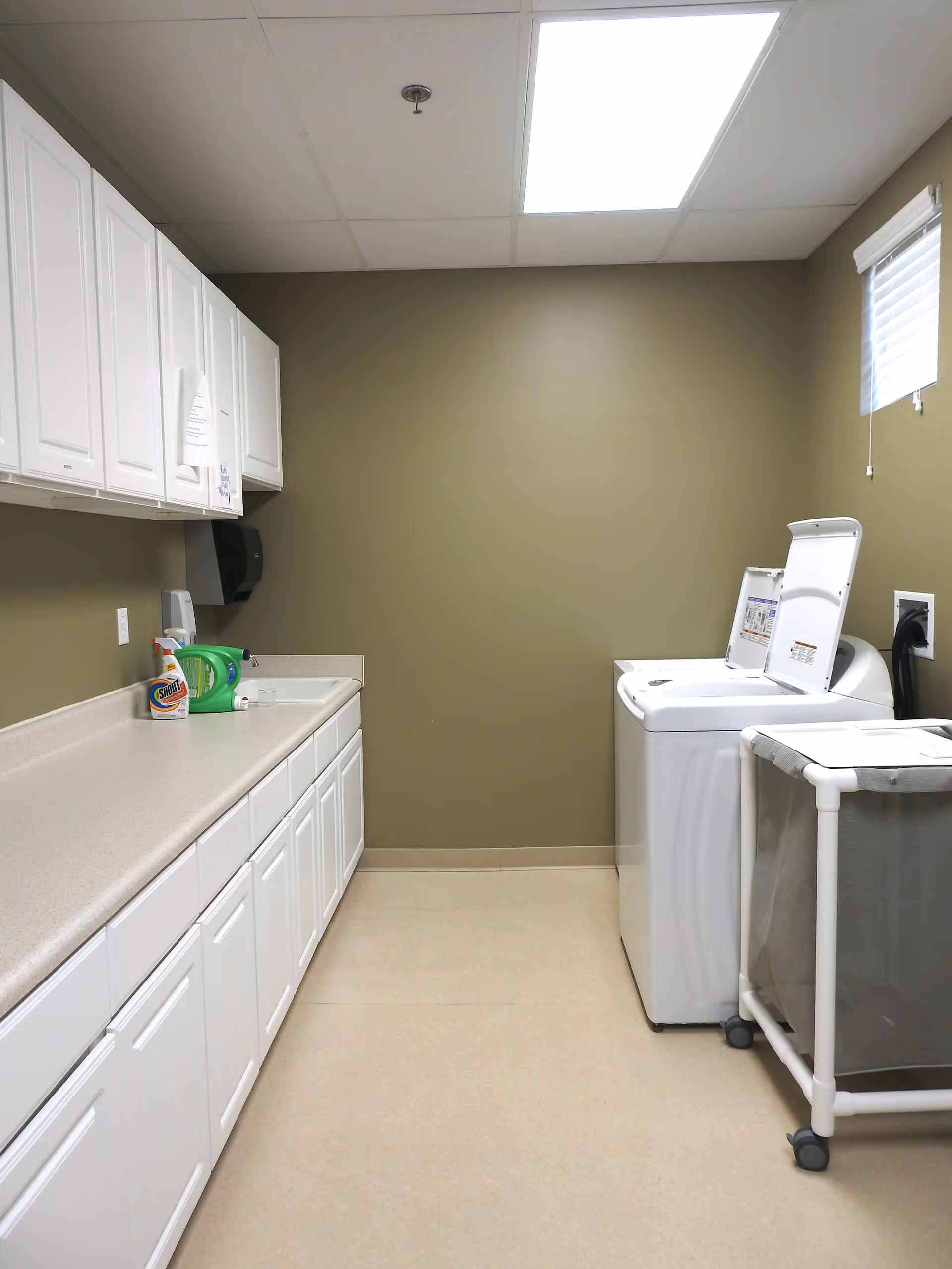 A clean laundry room with white cabinets, a countertop, a top-loading washing machine and a wheeled laundry cart.