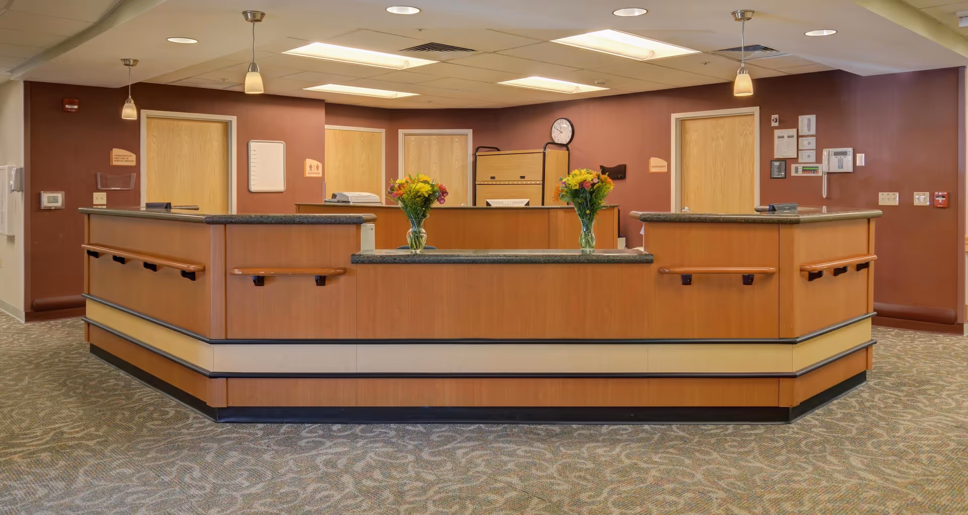 Reception desk area with a large wood-paneled counter, two vases of flowers, carpeted floor, and doors in the background.