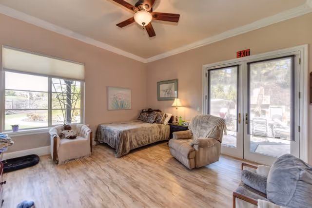 Bright senior living bedroom/lounge with a bed, recliner chairs, a ceiling fan, and glass doors leading to an outdoor patio.