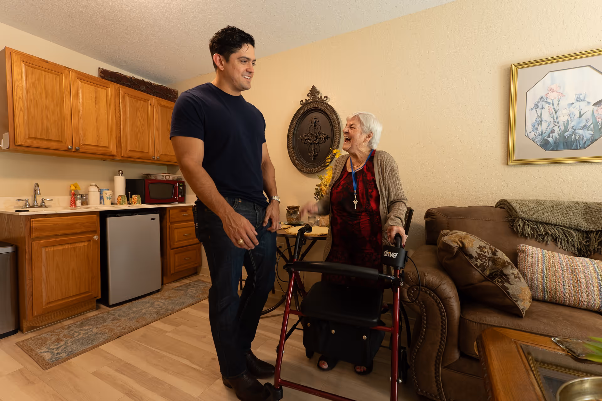 An elderly woman with a walker is smiling and talking with a younger man in a cozy living area that includes a small kitchen with wooden cabinets, a mini fridge, and a microwave. The room also has a brown couch with pillows and a framed floral picture on the wall.
